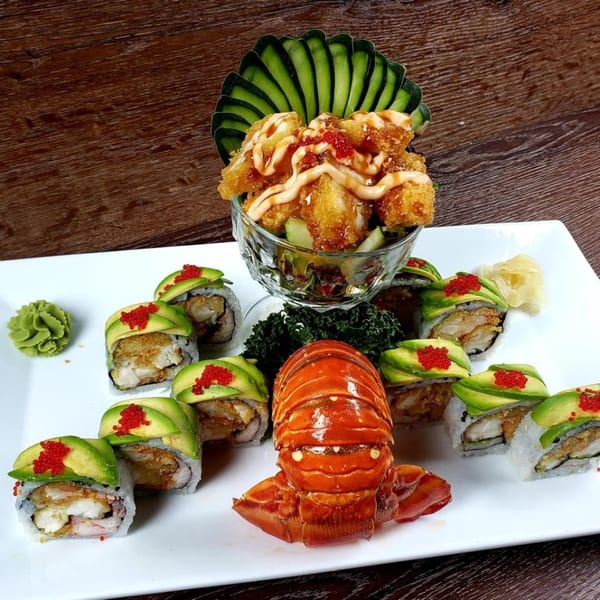 Lobster tail, avocado sushi rolls with red roe, and fried shrimp with sauce on a white plate.