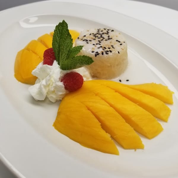Thai mango sticky rice dessert with whipped cream, raspberries, and mint on a white plate.